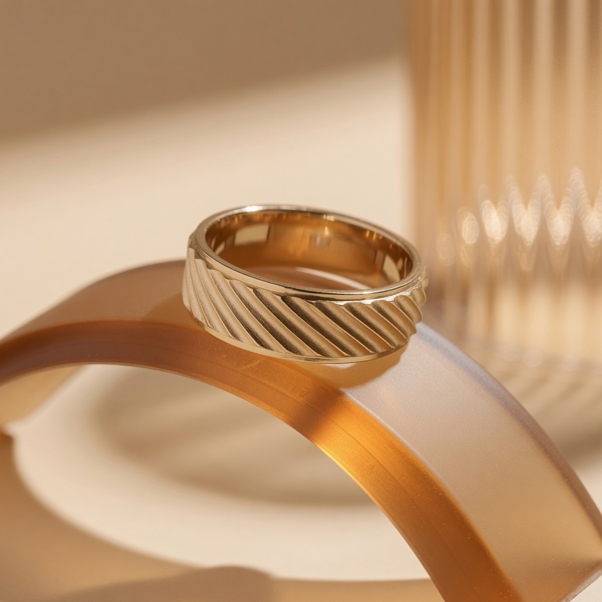 The Furrow: a diagonal twill-ridge solid gold wedding band, photographed against warm amber glass